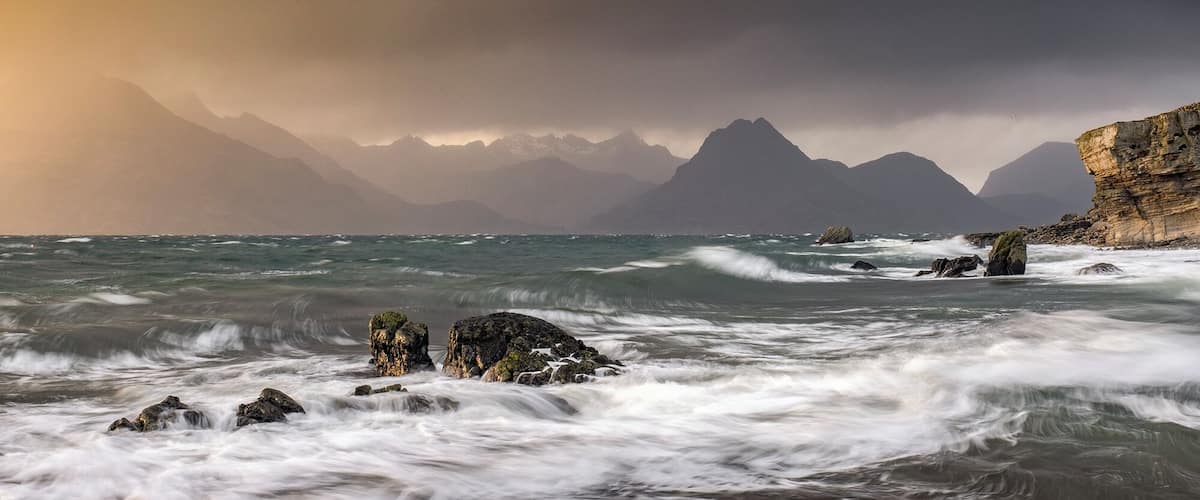 Una costa escarpada con olas turbulentas chocando contra rocas y acantilados, bajo un cielo dramático con montañas al fondo.
