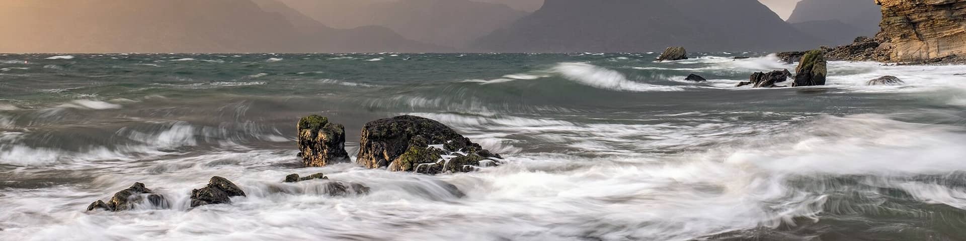 A rugged coastline with turbulent waves crashing against rocks and cliffs, under a dramatic sky with mountains in the background.