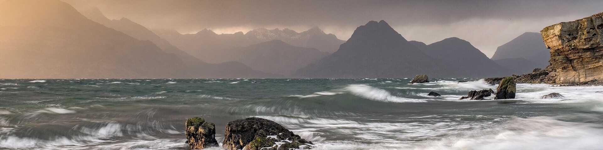 Una costa escarpada con olas turbulentas chocando contra rocas y acantilados, bajo un cielo dramático con montañas al fondo.