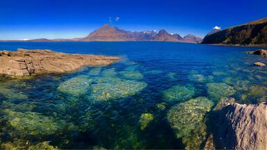 A clear coastal landscape with rocky shores, a calm sea, and distant mountains under a blue sky.