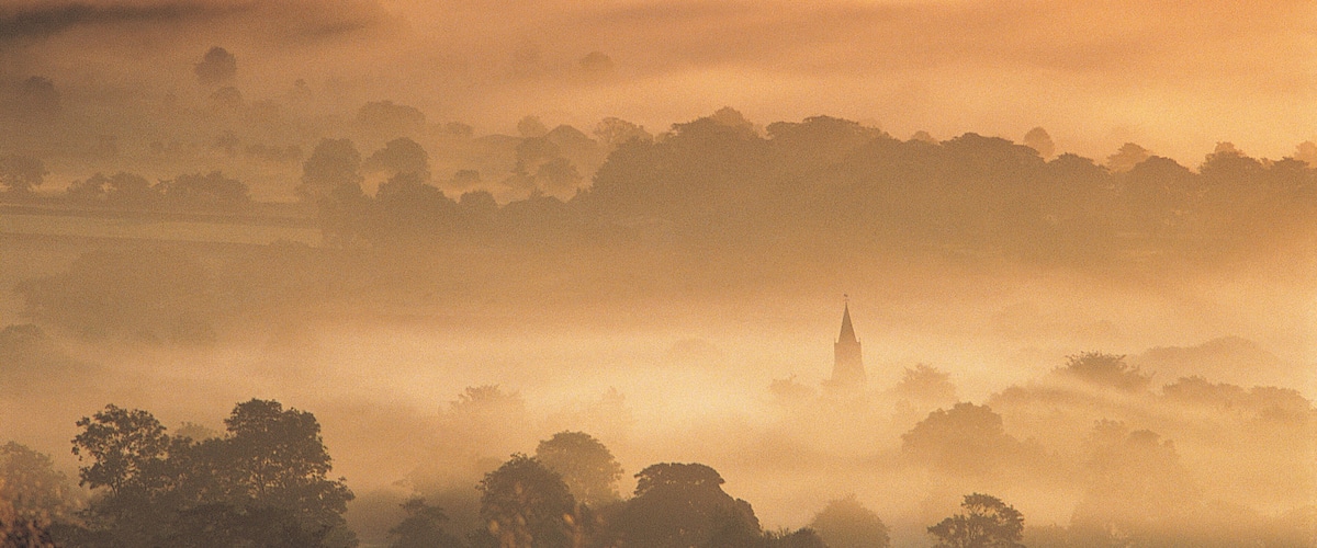 A misty landscape with rolling hills and a church spire in the distance.