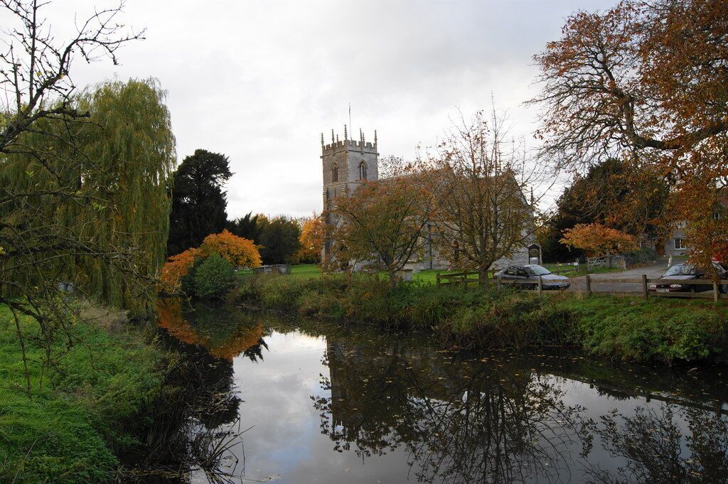 A calm river with autumn foliage, a historic tower, and a park with trees and a car.