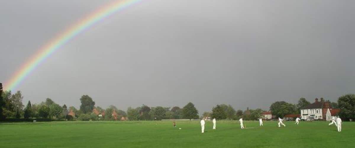 A cricket field with players, a rainbow, and houses in the background.