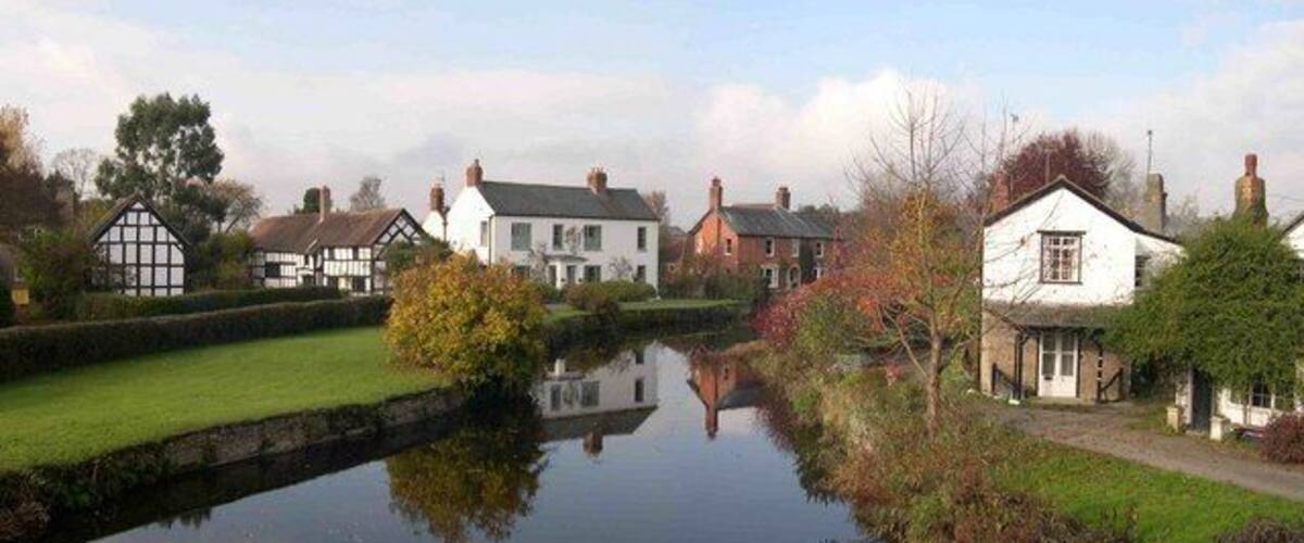 A calm river with houses on one side and a grassy area with trees on the other.