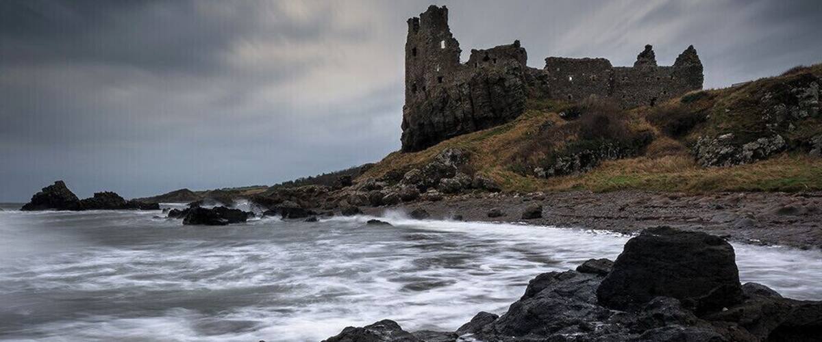 Paisaje costero con un castillo en ruinas y un mar agitado.