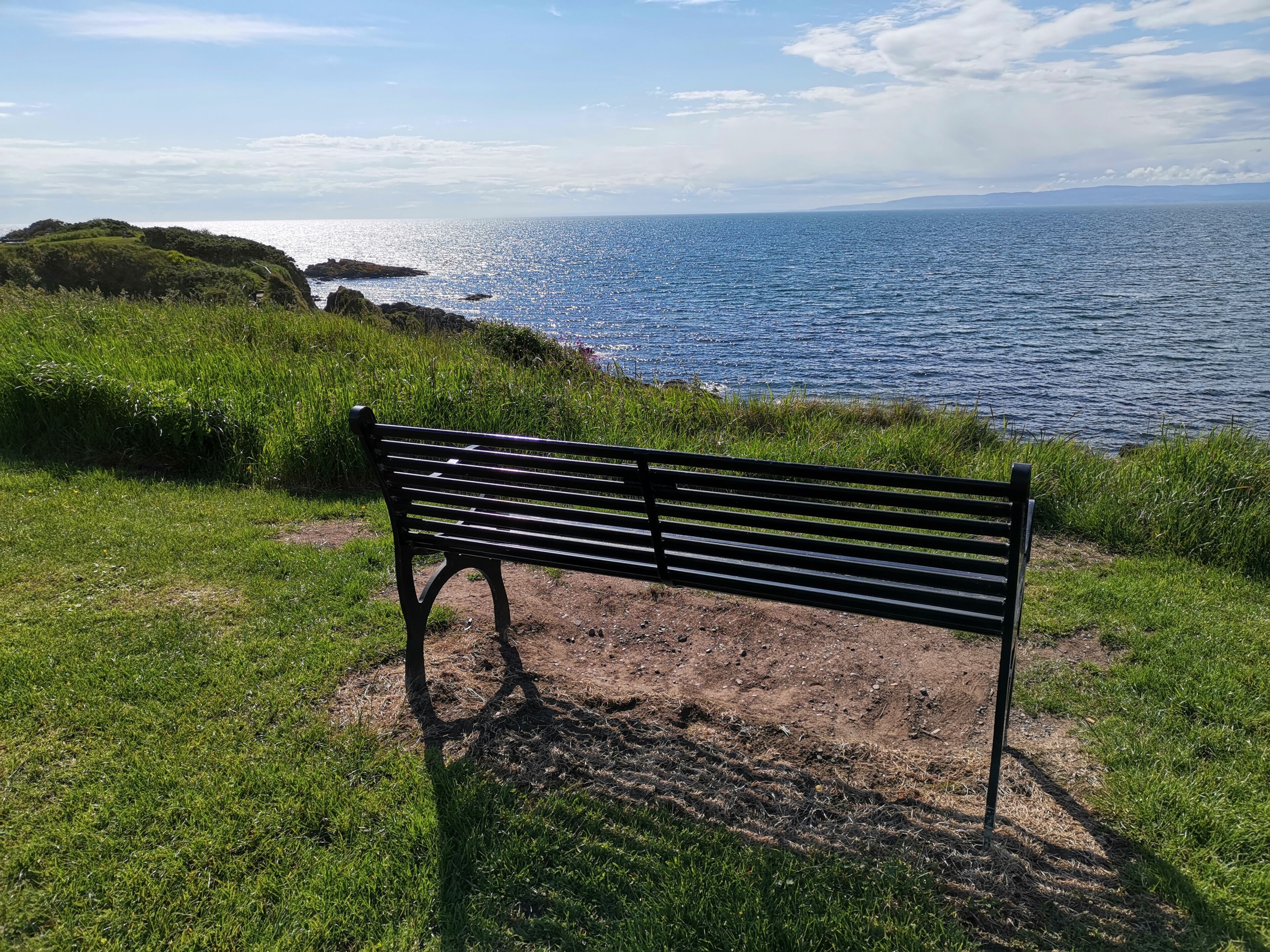 A bench overlooking a coastal landscape with a view of the sea and distant hills.