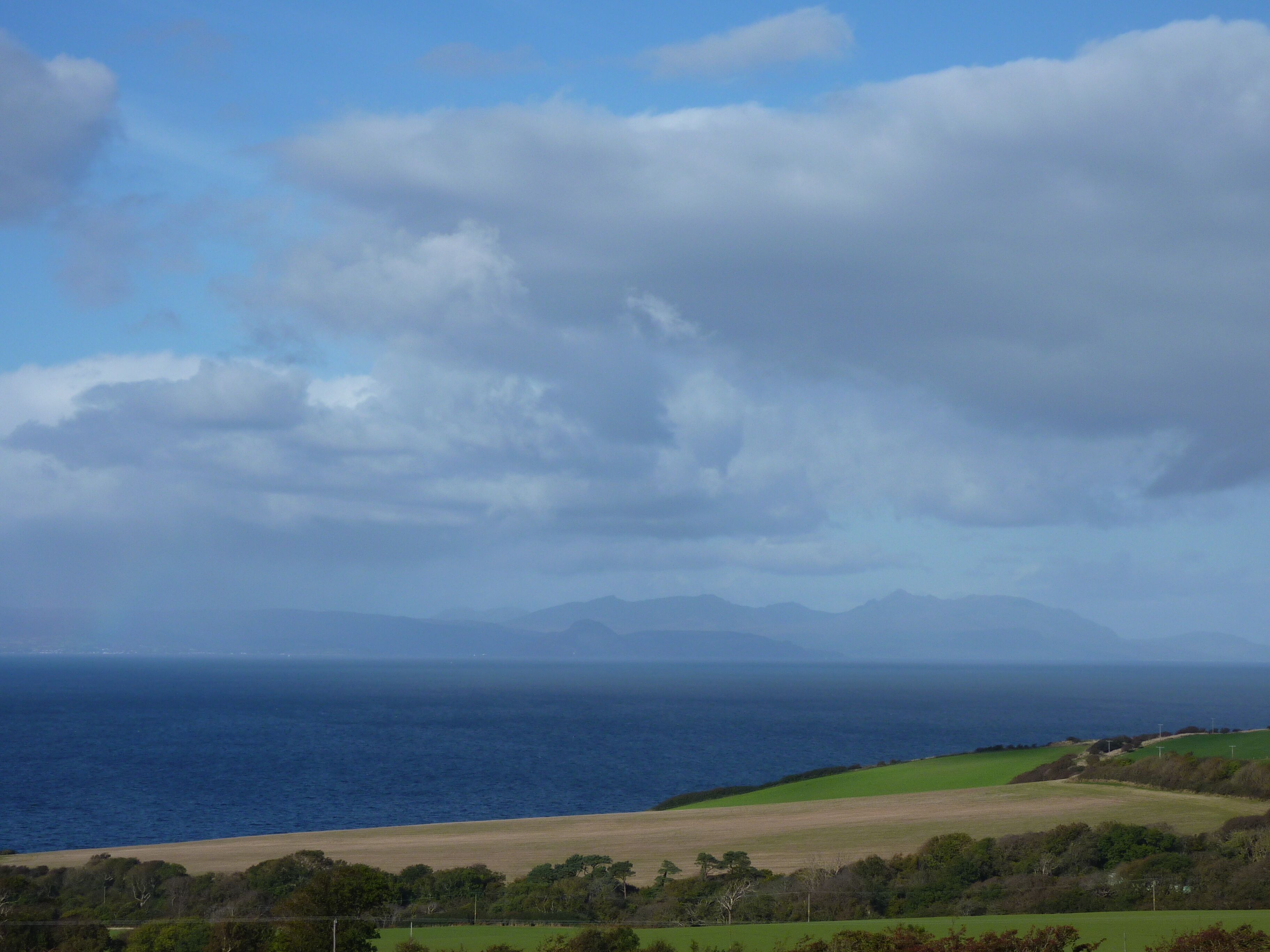 A coastal landscape with a view of the sea, green fields, and distant mountains under a cloudy sky.