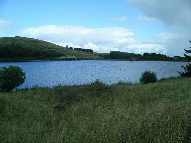 Un lago rodeado de colinas cubiertas de hierba y árboles bajo un cielo parcialmente nublado.