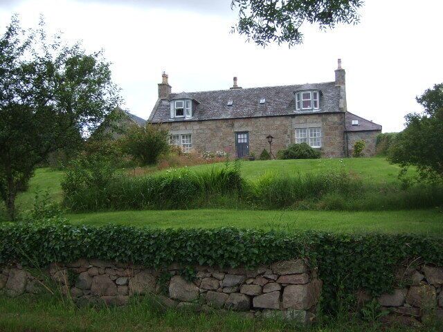 A stone house with a sloped roof, surrounded by a stone wall and lush greenery.