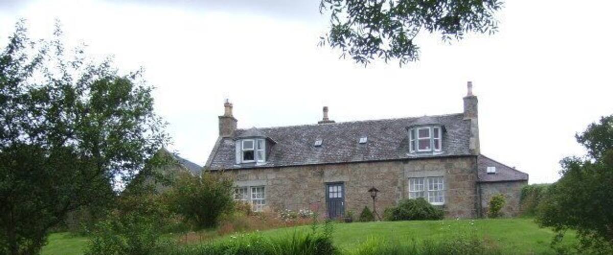 A stone house with a sloped roof, surrounded by a stone wall and lush greenery.