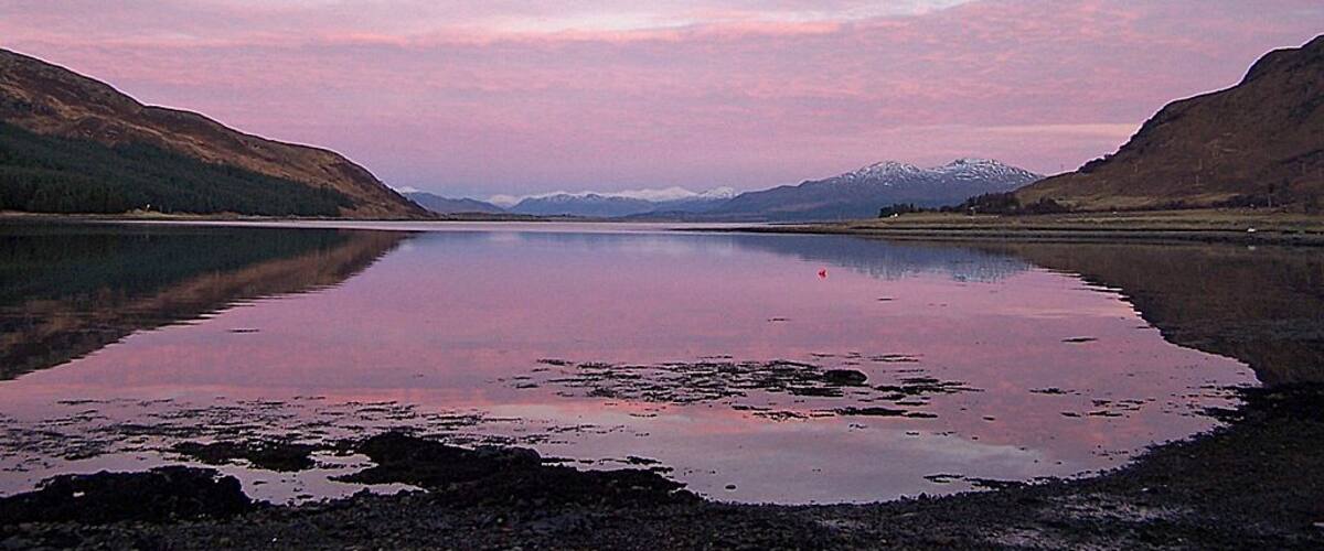 A calm lake with mountains in the background and a pink sky.
