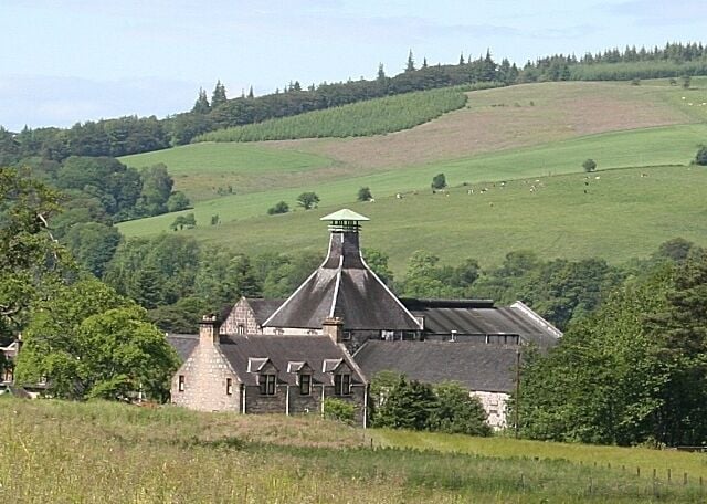 A large stone building with a conical roof, surrounded by lush green fields and rolling hills.