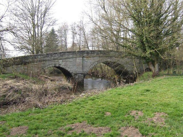 A stone bridge over a river with grassy banks and trees in the background.