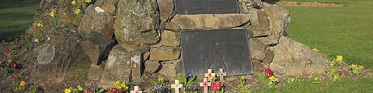 Une statue d’un soldat, posée sur un socle rocheux et agrémentée d’une plaque commémorative, le tout entouré de fleurs.