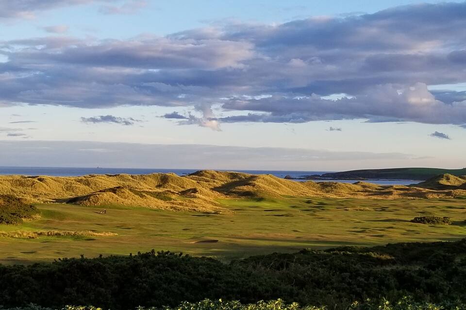 Paisagem costeira com dunas ondulantes, um corpo d'água ao longe e céu claro.