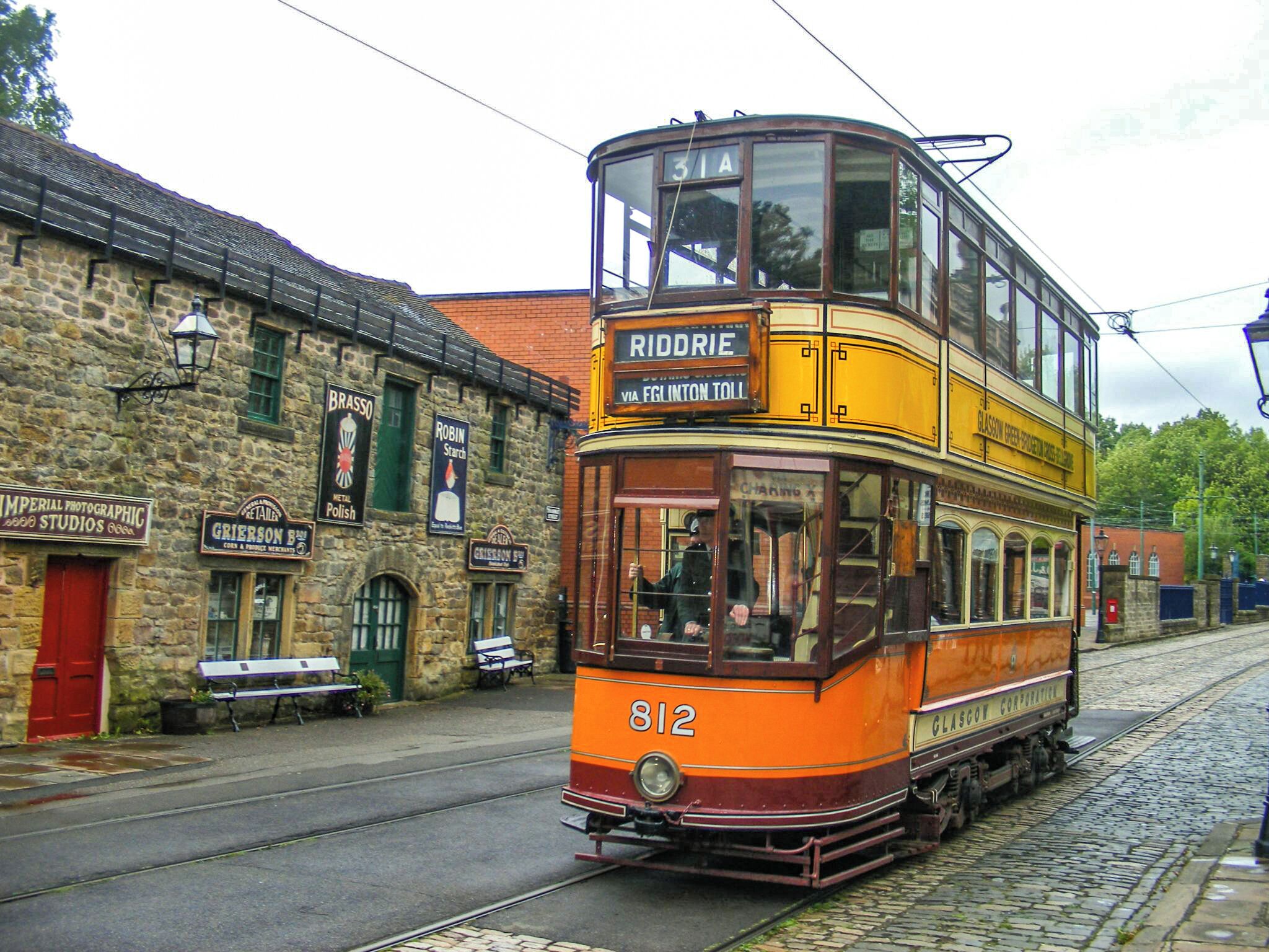 A vintage orange double-decker tram with the number 812, labeled 'Riddrie' and 'Via Fglinton Toll', traveling on a cobblestone street past historic buildings with signs for 'Brasso', 'Robin Starch', and 'Imperial Photographic Studios'.