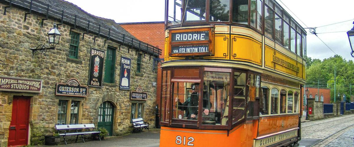 A vintage orange double-decker tram with the number 812, labeled 'Riddrie' and 'Via Fglinton Toll', traveling on a cobblestone street past historic buildings with signs for 'Brasso', 'Robin Starch', and 'Imperial Photographic Studios'.