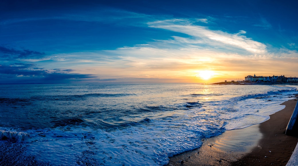 Atardecer costero con olas, una playa arenosa y una costa distante con edificios.