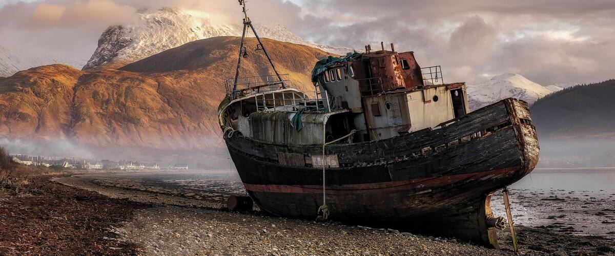 A weathered boat stranded on a rocky shore with mountains in the background.