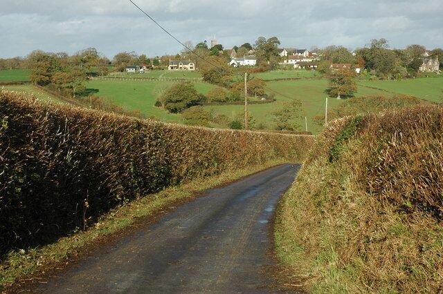 Road approaching Down St Mary The village of Down St Mary and its church tower can be seen on the hill in the background.