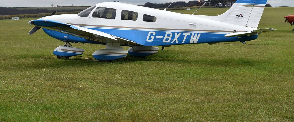 A small white and blue Piper PA-28 aircraft with the registration G-BXTW parked on grass.
