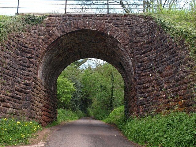 Railway bridge, Nethercott. A view of 35701 from the other side, looking up the lane to Dull Cross. The bridge is listed; it dates from about 1860, and is built of "red sandstone and conglomorate, coursed, squared, dressed masonry, ashlar archivolt of arch. Single arch with splayed base, rusticated voussoirs, no parapet. Curved walls abutting bank ... Much of the sandstone came from the quarry situated Southwest side ... One of the most impressive railway bridges on the West Somerset Railway". http://www.imagesofengland.org.uk/Details/default.aspx?pid=2&id=270290