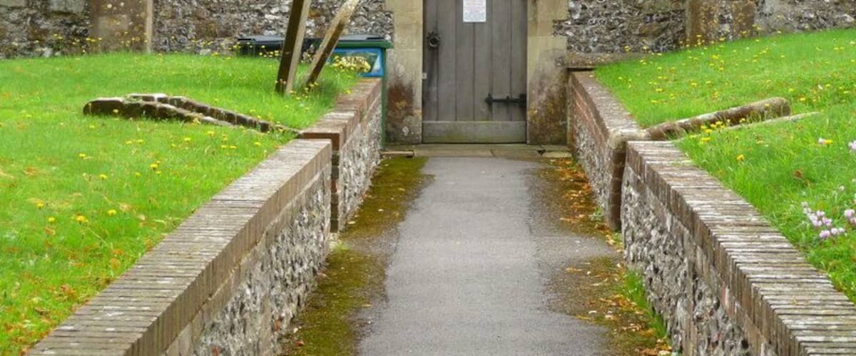 A stone building with a wooden door, a narrow pathway, and a grassy area with flowers.