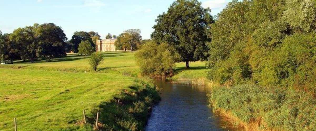A river flowing through a grassy field with trees and a building in the background.