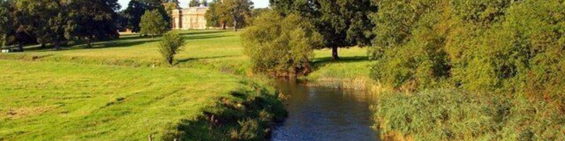 A river flowing through a grassy field with trees and a building in the background.