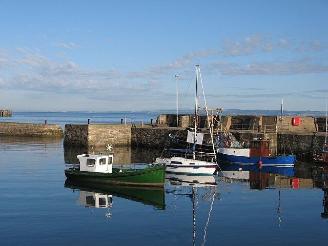 A calm harbor with several boats docked, a stone wall, and a clear sky.
