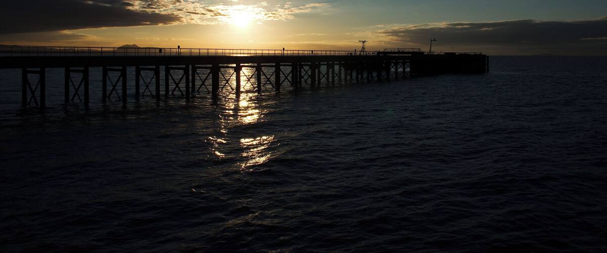 A pier extending into calm waters at sunset with the sun low on the horizon.