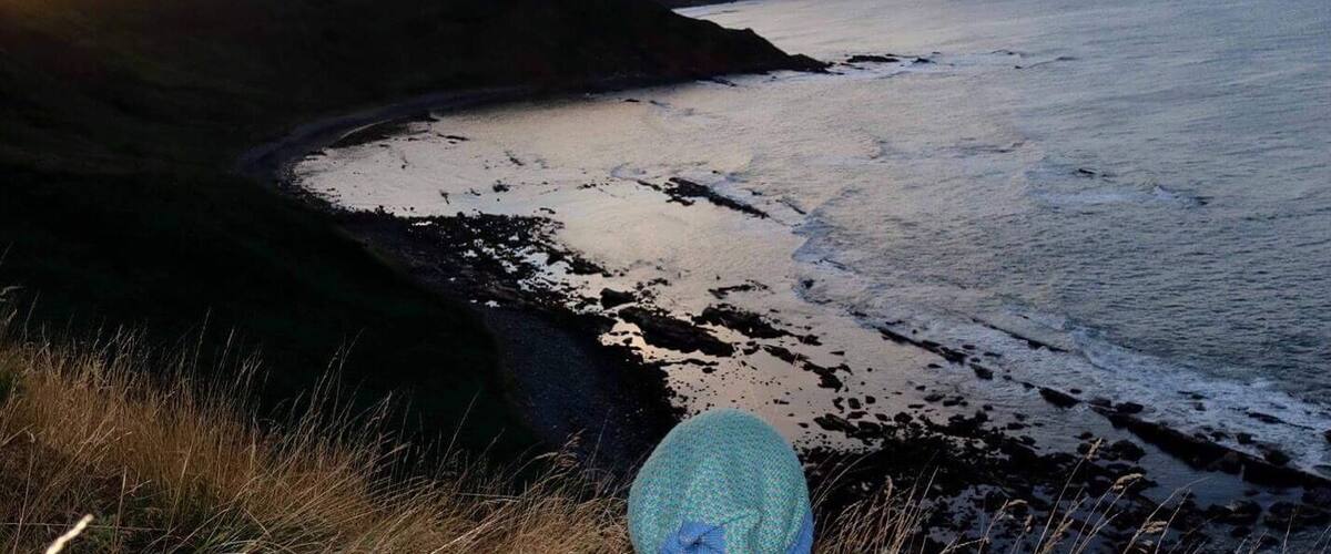 A coastal landscape with a person sitting on a grassy hill overlooking the sea during sunset.