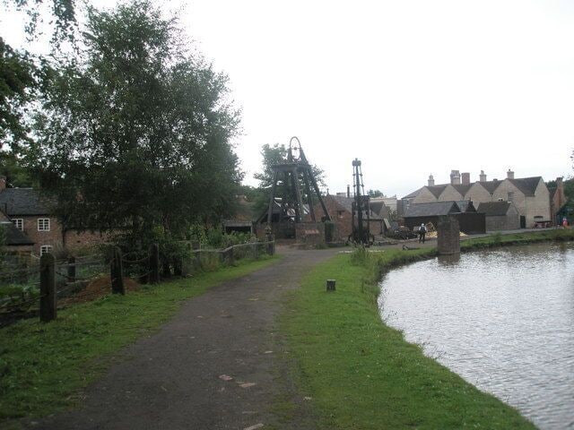 A narrow path beside a canal, with a water wheel and buildings in the background.