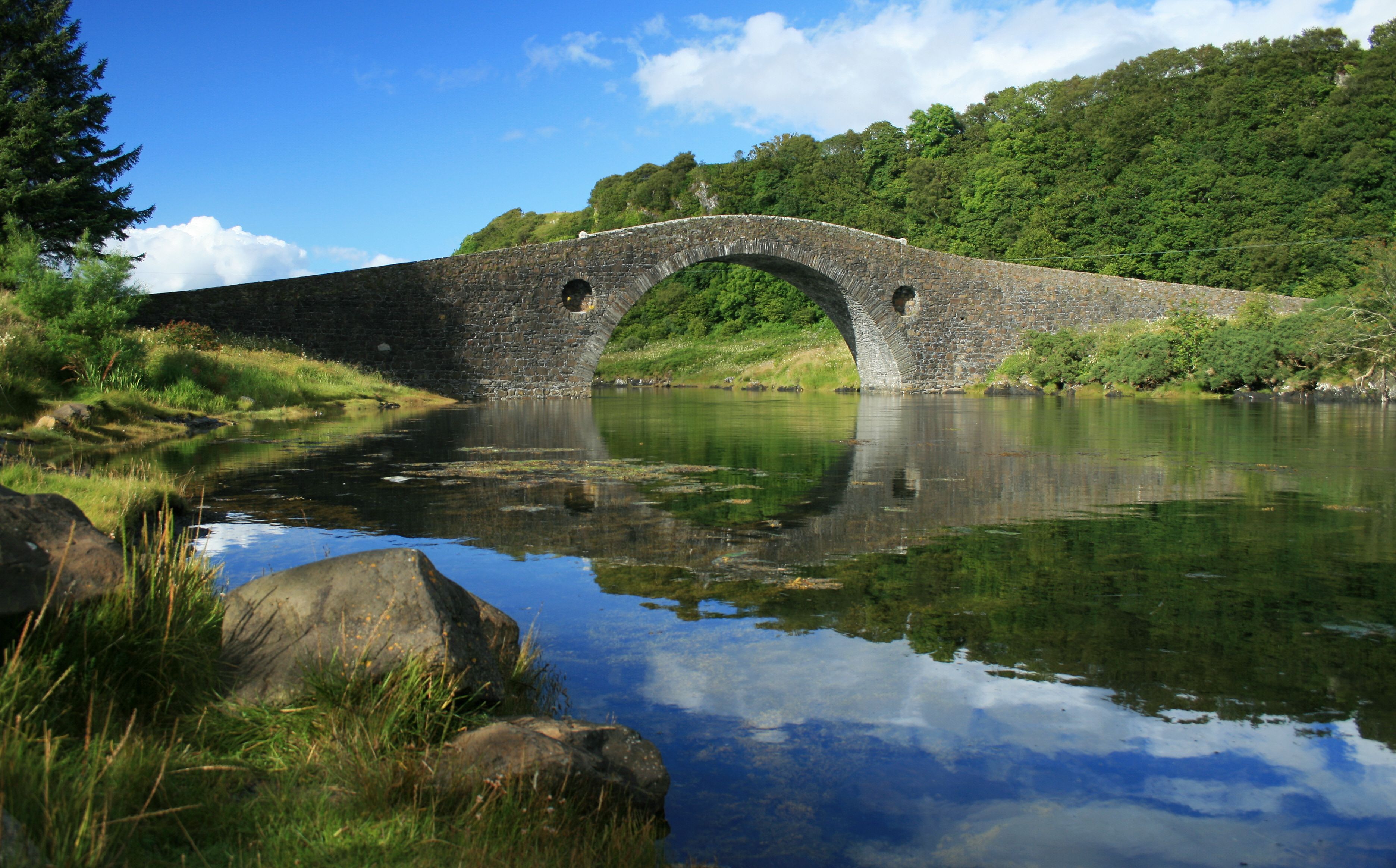 A stone bridge over a calm river with lush greenery and a clear sky.
