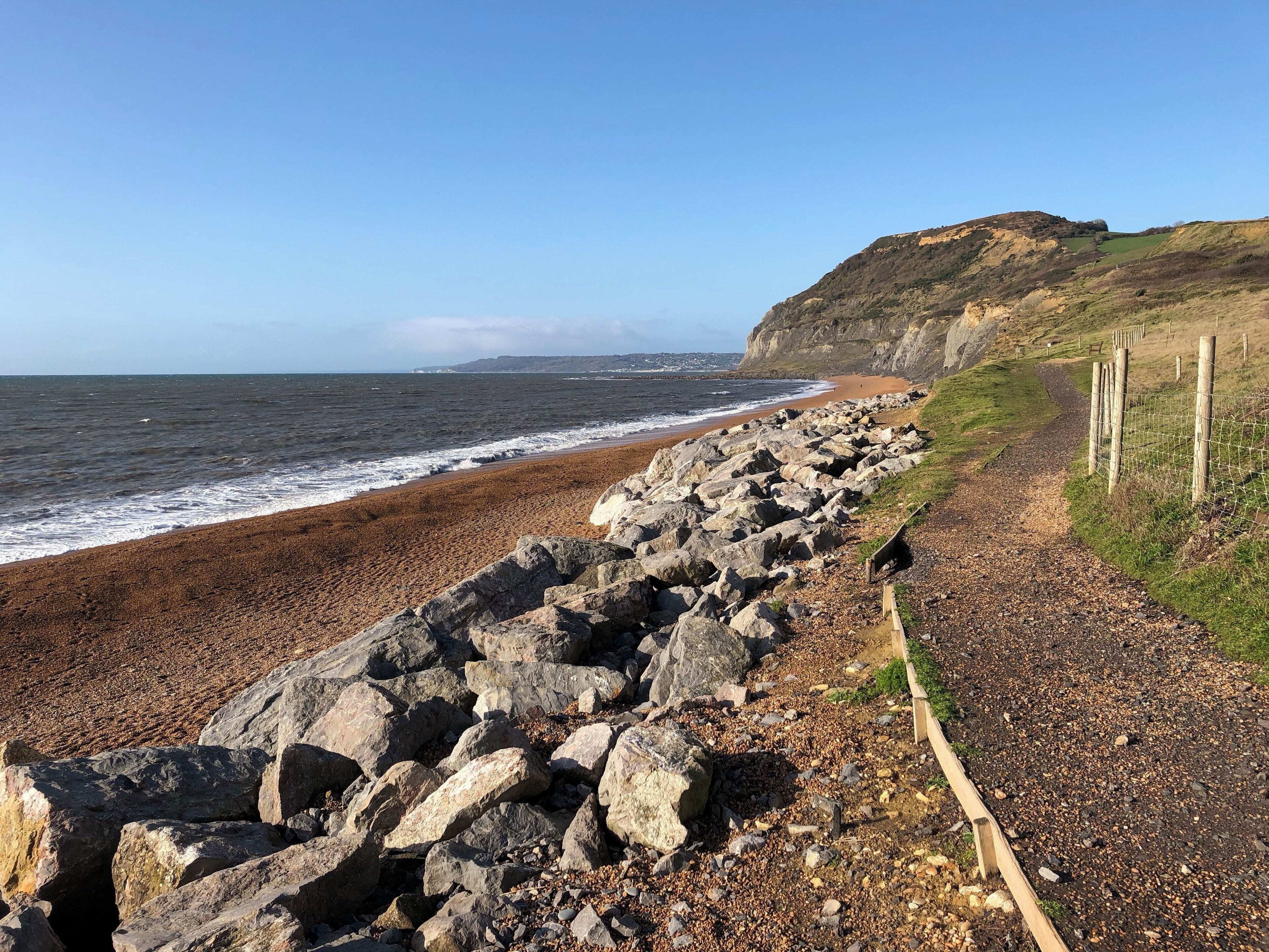 A rocky beach with waves crashing onto the shore, a grassy path, and a cliff in the background.
