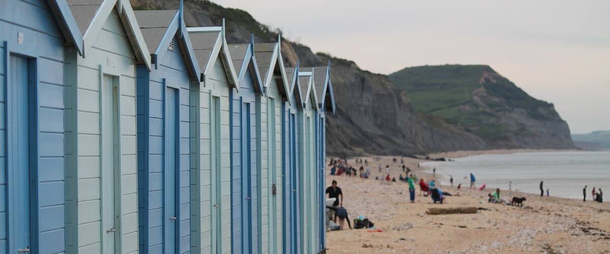 A row of blue beach huts along a sandy beach with people in the background and cliffs in the distance.