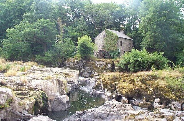 Una casa de piedra cubierta de hiedra, una orilla rocosa del río y árboles frondosos.