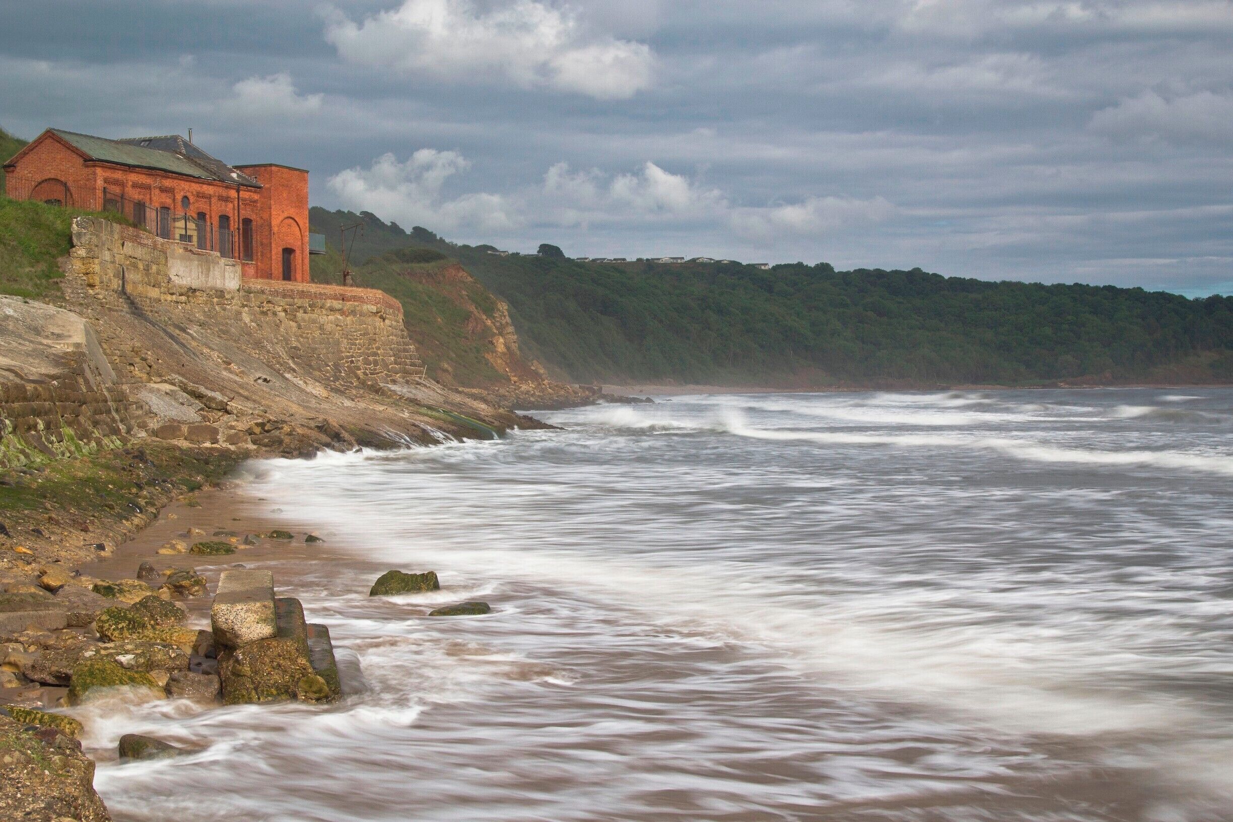 A coastal scene with a red brick building on a cliff, waves crashing onto the shore, and a forested hillside in the background.