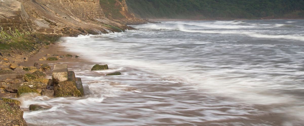 A coastal scene with a red brick building on a cliff, waves crashing onto the shore, and a forested hillside in the background.
