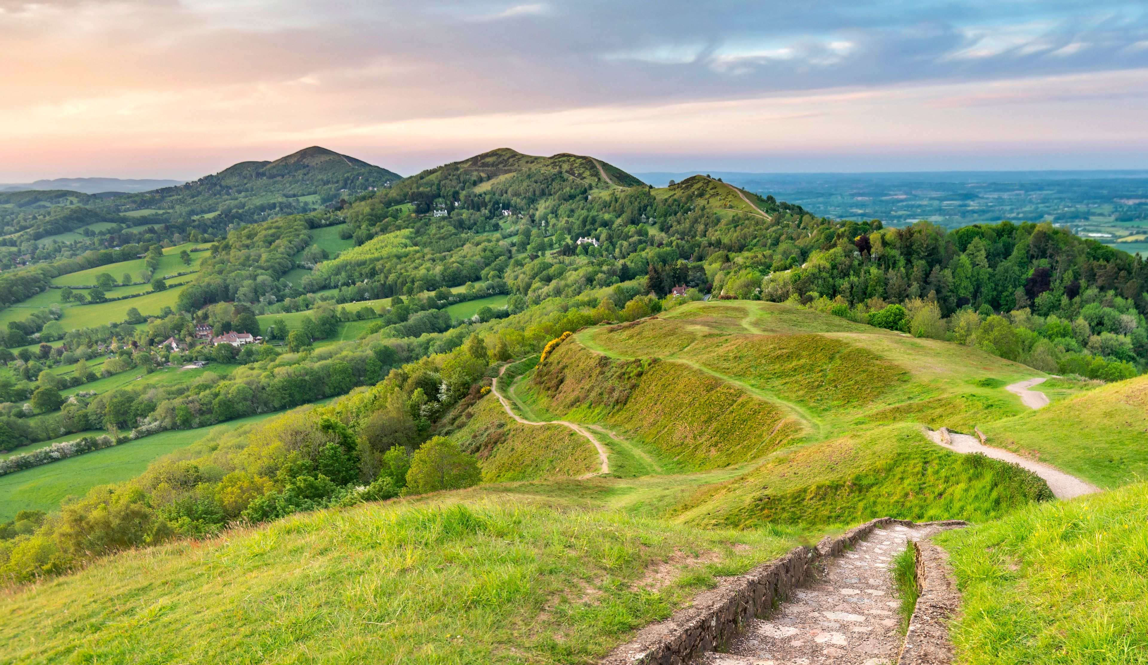 A lush green valley with rolling hills, a winding path, and a distant view of a house on a hill.