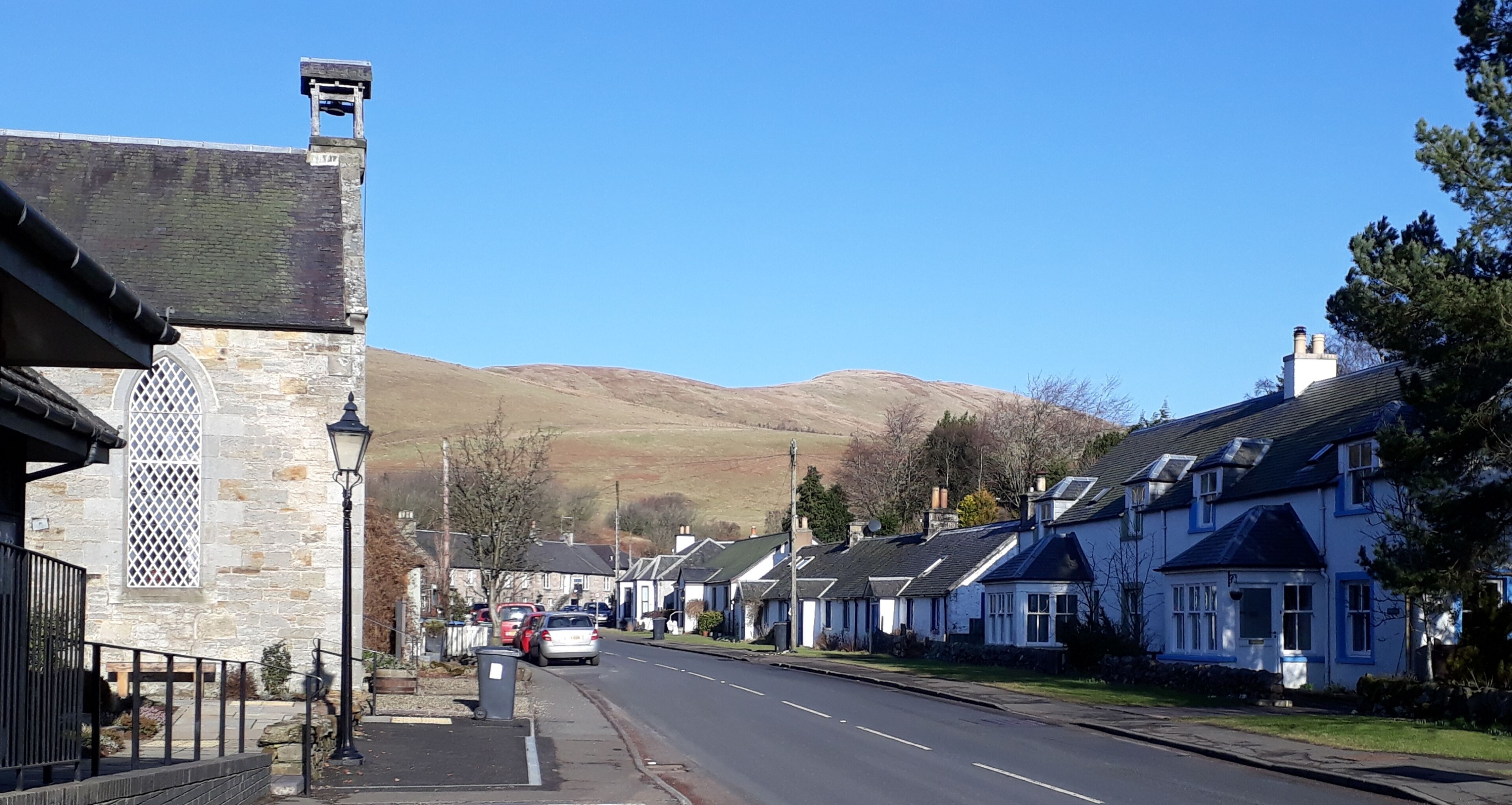 A street with stone buildings, parked cars, and a mountain in the background.