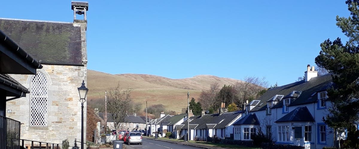 A street with stone buildings, parked cars, and a mountain in the background.
