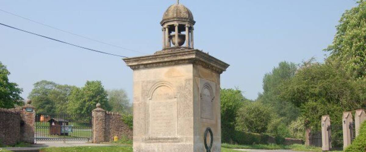 A stone monument with a dome and bell tower, surrounded by grass and a road.