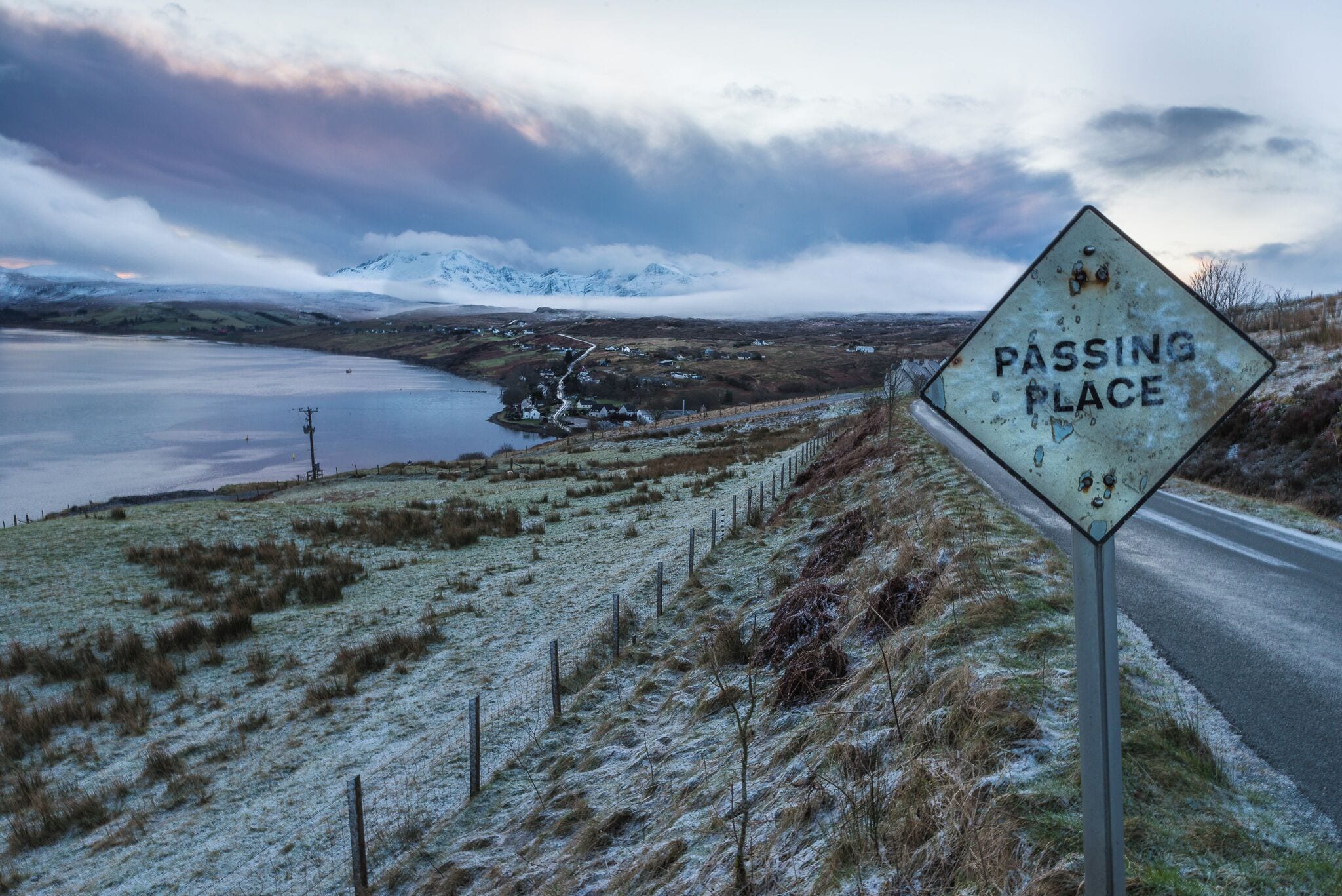 A 'Passing Place' sign on a rural road with a lake and mountains in the background.