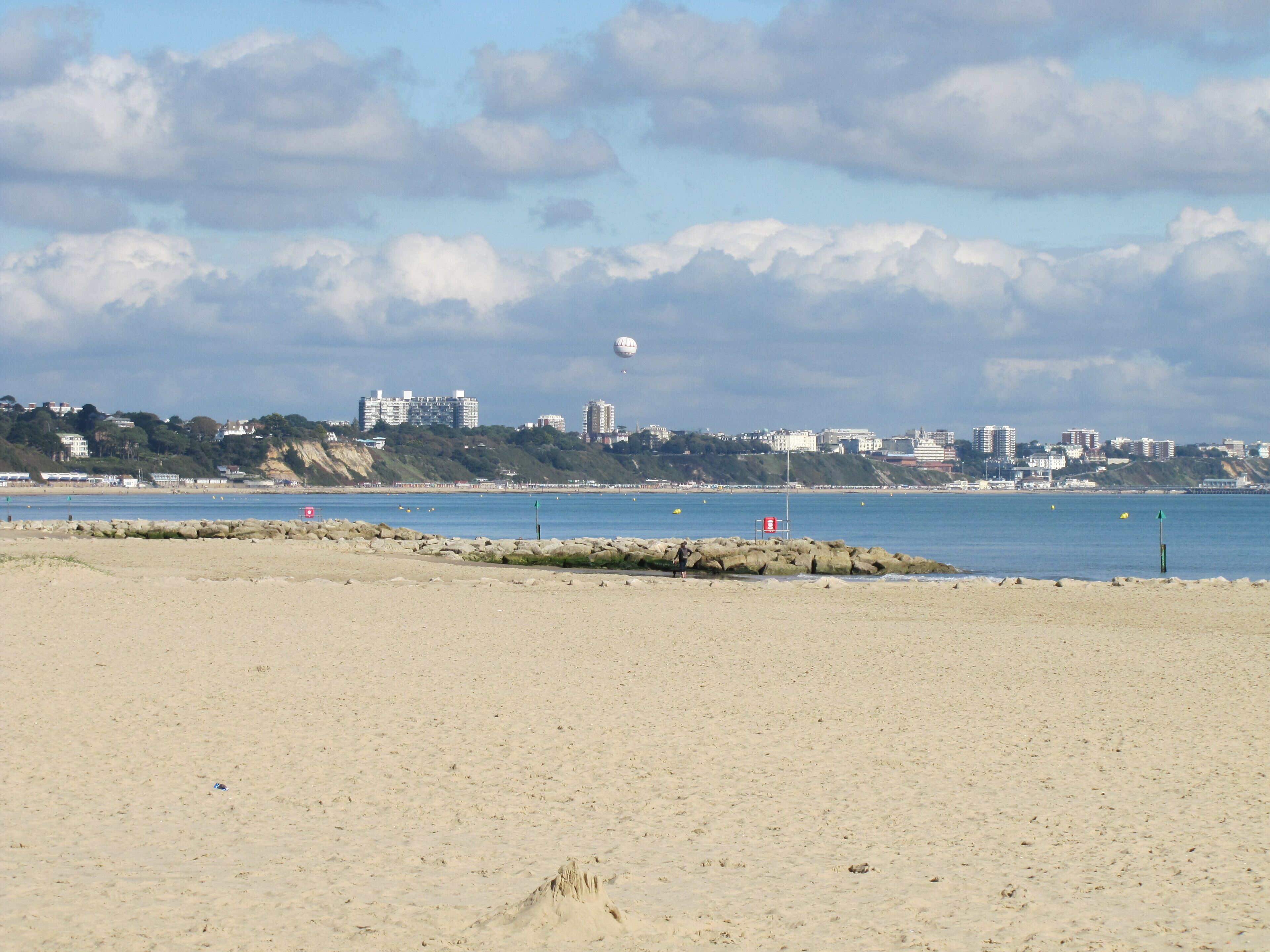 A sandy beach with a rocky breakwater, calm sea, and a cityscape in the background.