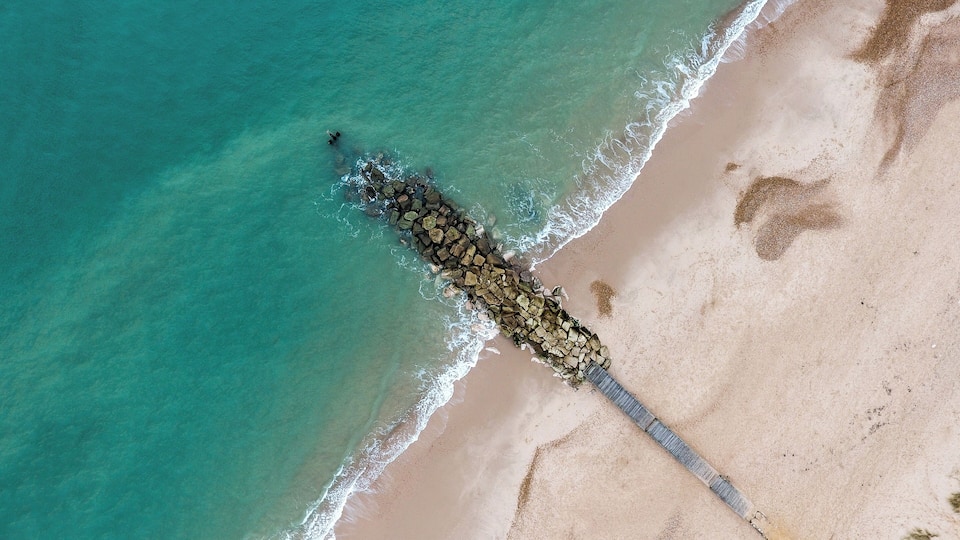 An aerial view of a beach with clear turquoise water, a sandy shore, and a long, narrow breakwater extending into the sea.