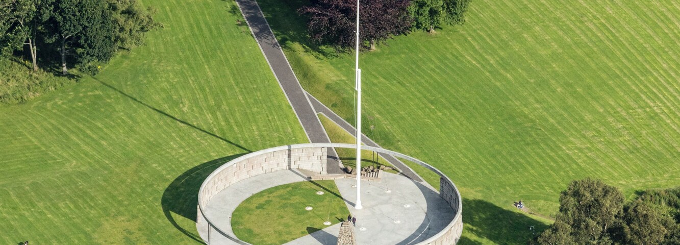 An aerial view of a circular monument with a flagpole, surrounded by a well-maintained lawn and a road.