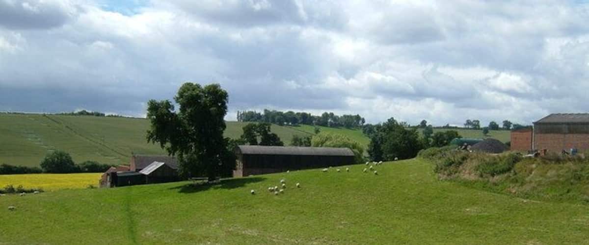 A rural landscape with a barn, sheep, and rolling green fields under a partly cloudy sky.