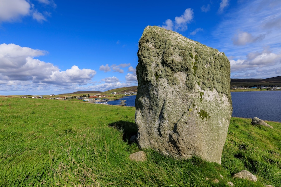 Una gran piedra vertical en un campo cubierto de hierba, con un lago y casas al fondo.