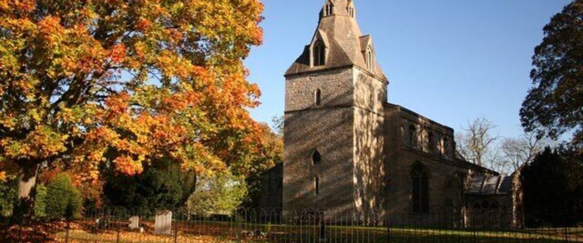 A church with a tall steeple and a cross on top, surrounded by autumn trees and a fenced area.